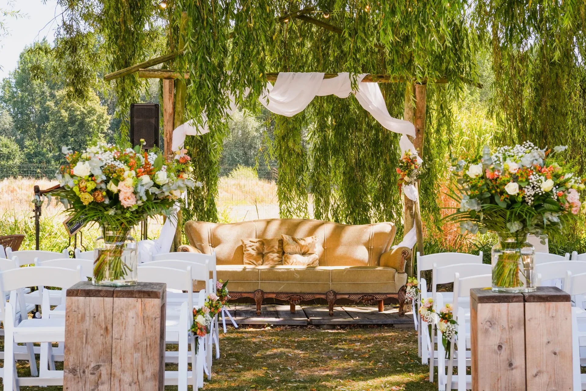 Ceremonie-opstelling in de tuin met witte stoelen, bloemen en een gouden sofa onder een treurwilg
