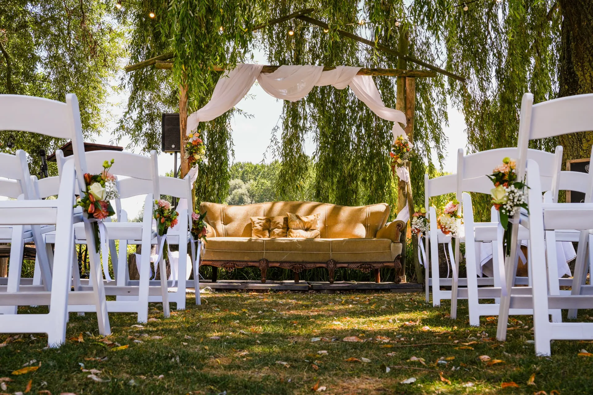 Ceremoniegangpad met witte stoelen richting de sofa en ceremonieboog onder de treurwilg